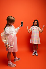 full length of happy girl showing win gesture near friend taking photo on mobile phone on orange background