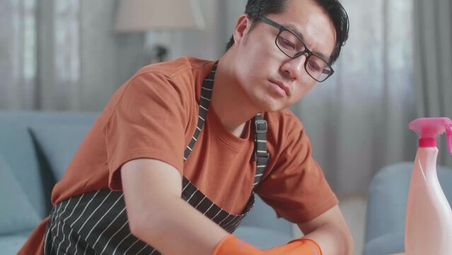 Close Up Of Tired Asian Male Housekeeper With An Apron Cleaning The Table By The Spray At Home
