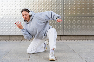 Young woman with headphones listening to urban music and dressed in white pants and gray hoodie dancing in front of metallic background urban dance