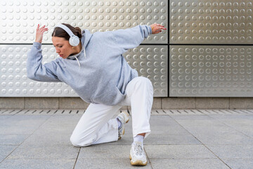 Young woman with headphones listening to urban music and dressed in white pants and gray hoodie dancing in front of metallic background urban dance