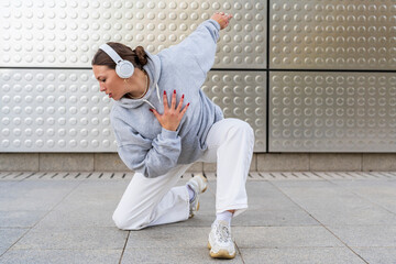 Young woman with headphones listening to urban music and dressed in white pants and gray hoodie dancing in front of metallic background urban dance