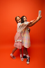 full length of girl pointing with finger near friend taking selfie on mobile phone on orange background