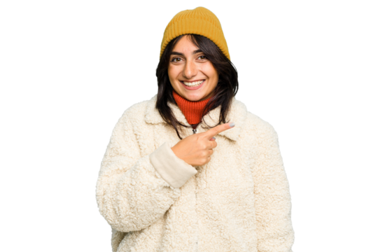 Young Indian woman wearing winter jacket and a wool cap isolated smiling and pointing aside, showing something at blank space. - Powered by Adobe