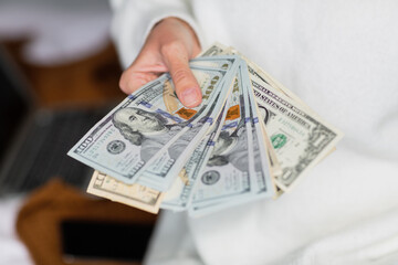 Young caucasian woman holding hands cash money dollars bills. Person counting money at home on bed, next lying laptop and mobile phone.