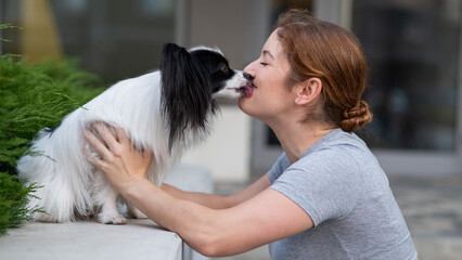 Caucasian red-haired woman kissing pappilion dog outdoors. Black and white continental spaniel.