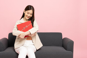 Happy beautiful Asian woman smile hugging red gift box and sitting on sofa isolated on pink...