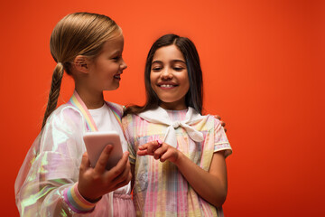 smiling girl pointing at blurred smartphone in hand of happy friend isolated on orange