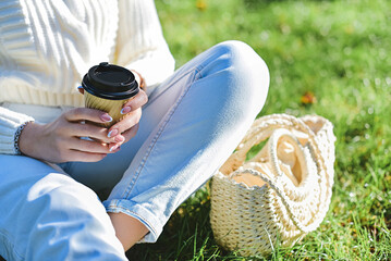 A woman in a white sweater and blue jeans sits on the grass and holds a glass of coffee in her hands.