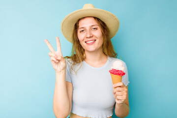 Young caucasian woman holding an ice cream isolated a blue background joyful and carefree showing a peace symbol with fingers.