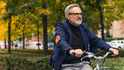 Millennial happy bearded grey-haired mature man with eyeglasses enjoying warm weather during the fall season while riding a bicycle around trees with yellow leaves. High quality photo