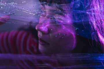 Close up face of a young, caucasian girl with long dark hair looking aside. Purple led lights, a photo through plastic wrap. High quality photo