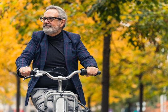 Stylish and happy bearded grey-haired mature man enjoying warm weather during the fall season while riding a bicycle around trees with yellow leaves. High quality photo