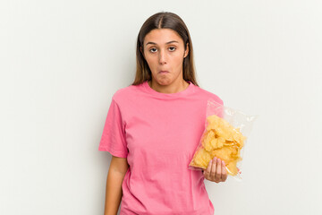 Young Indian woman holding crips isolated on white background shrugs shoulders and open eyes...