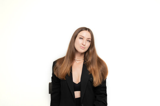 A beautiful long-haired girl dressed in a black jacket, bra and trousers sits on a chair in a photo studio on a white background