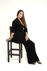 A beautiful long-haired girl dressed in a black jacket, bra and trousers sits on a chair in a photo studio on a white background