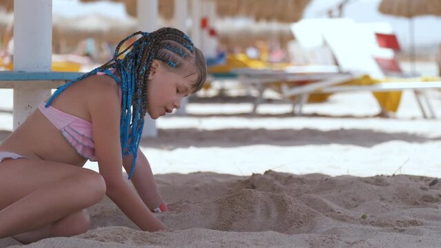 Pretty child girl in bikini swimsuit playing with sand in umbrella shadow during summer tropical vacation