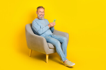 Fototapeta na wymiar Full size photo of cheerful man sit chair hold use telephone chatting isolated on yellow color background