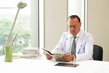 Senior doctor reading medical textbook at his Office Desktop, examining Medical Reports. Health care, Medical and Pharmacy Concept. Professional Doctor working at hospital.