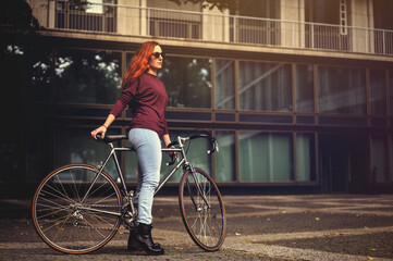 Fototapeta na wymiar beautiful red-haired woman with sun glasses, sexy tight blue jeans and black boots stands over top tube of a vintage road bike in dark urban scenery. selective focus. copy space for text