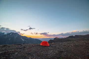 Scenic alpine landscape with tent at very high altitude with view to large mountains in orange dawn sky. Vivid orange tent with awesome view to high mountain range under cloudy sky in sunset colors.