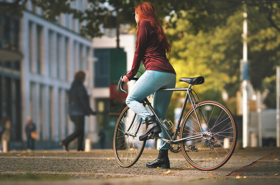 beautiful red-haired woman in black boots and burgundy top stands over top tube of a vintage road bike in city scenery. selective focus. view on back side in tight blue jeans. urban lifestyle concept