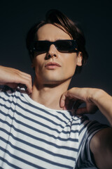 Trendy man in sunglasses touching striped t-shirt isolated on grey with lighting