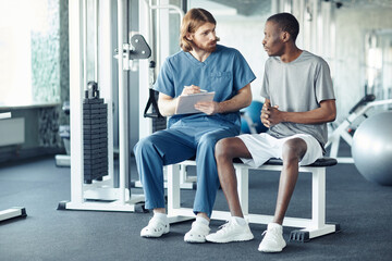 Young doctor asking questions to patient about his rehabilitation while they sitting in gym