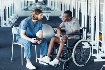 Young doctor in uniform discussing rehabilitation with patient while he sitting in wheelchair in gym