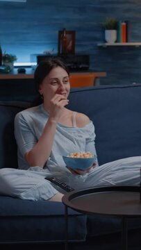 Vertical video: Relax woman in pajamas eating popcorn watching TV, enjoying mid-night entertainment show sitting on confortable couch. Young lady spending free time relaxing looking at television at