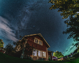 Milky Way above the Swedish traditional wooden red house at the right side of night scene in garden. Starry sky, green glowing clouds, green grass.