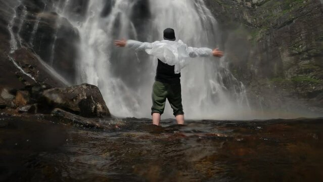 Happy man in a transparent raincoat stands with hands up at the foot of a waterfall in norway near a rock