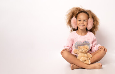 beautiful african american child wearing earmuffs hat hugging teddy bear and smiling at camera isolated on white