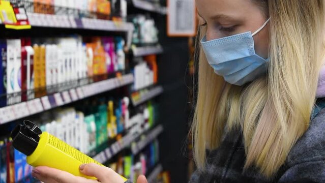 Young Girl Customer In Protective Mask Choosing Hair Cosmetic Products In Hygiene Department Of Mall. Woman Reading Composition On Label At Supermarket. Close Up Background Of Shopping Shelves