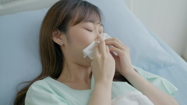 Asian woman sneezing in the hospital room. Sick woman has the flu or cold symptoms and blows her nose. Close up of Asian woman sneezing into tissue in hospital.