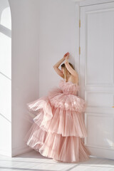 Naklejka premium A girl in a beautiful lush long dress of the color of a dusty rose stands near a light wall. Vintage bridal glamor. Fashion shooting in a wedding or evening dress