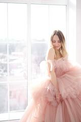 A girl in a beautiful lush long dress of dusty rose color stands near an open window. Vintage bridal glamor. Fashion shooting in a wedding or evening dress