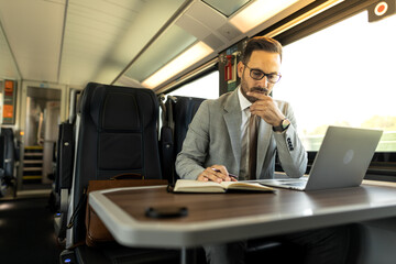 Young business man traveling to work by train. 
Working while traveling, using laptop, mobile phone and notebook
Thinking about new goals, noting, planning work