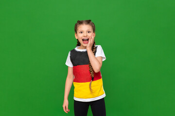 A little girl in a T-shirt with a German flag smiles in surprise on a green isolated background. German language courses for schoolchildren.