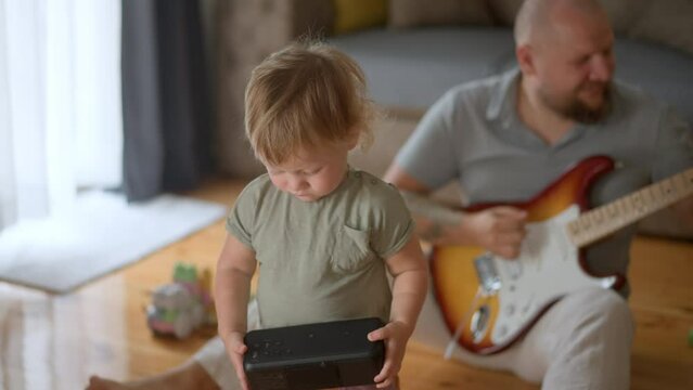 Smiling young father holds his little cute baby-boy on his knees and tries to teach her to play the guitar, cheerful toddler claps the guitar as if she playing it. Modern family, happy memories. Slow