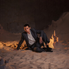 Financier and sand castles, the concept of unprofitable investments. Economic crisis and recession. A young man in a business suit is sitting on the sand next to a sand castle