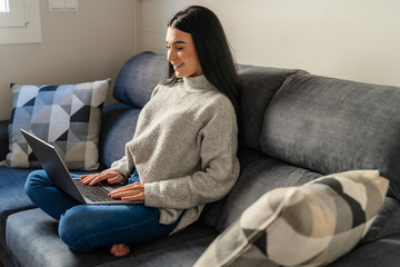 Young woman with long dark hair sitting on the sofa by a window working with the laptop on her legs