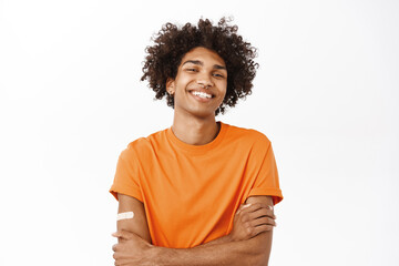 Portrait of smiling queer guy with vaccine shot, patch on arm from vaccination, covid-19 pandemic concept, standing over white background