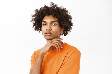 Fototapeta na wymiar Portrait of hispanic guy with curly hair, queer man with nose ring, looking confident at camera, standing in orange t-shirt over white background