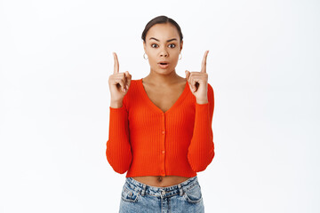 Portrait of surprised woman pointing, looking up, saying wow, gasping impressed, standing over studio background