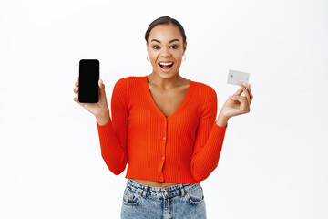 Happy young woman shows her mobile phone screen and credit card, smiling with excitement, stands...