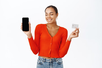 Happy young woman shows her mobile phone screen and credit card, smiling with excitement, stands over white background