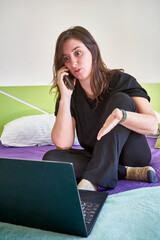 Young woman sitting on a bed with a laptop talking on a phone while explaining something