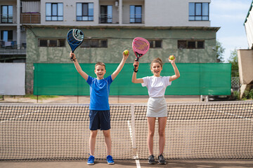 Kids and sports concept. Portrait of smiling boy and girl posing outdoor on padel court with rackets and tennis balls