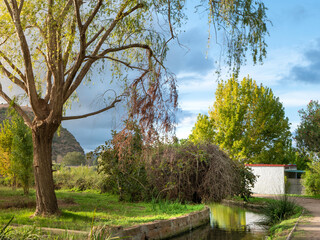 Beautiful landscape of a small park with trees, a building, a small canal and a mountain against a cloudy sky in the sunset rays of the sun