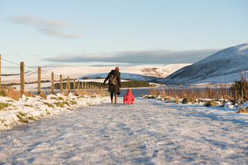 Low angle view of woman wearing a winter jacket and a woolly hat pulls a sledge with her daughter...